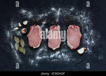 Top view of three fresh raw pork chops placed on a dark textured surface with scattered flour, peppercorns, garlic cloves, bay leaves, onion, and herb Stock Photo