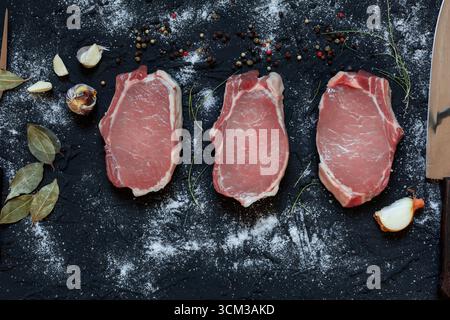 Top view of three raw pork chops placed on a dark textured surface surrounded by a chef’s knife, carving fork, garlic cloves, bay leaves, onion, peppe Stock Photo