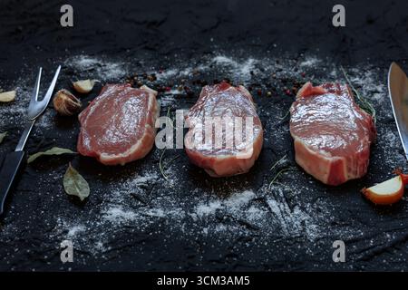 Top view of three raw pork chops placed on a dark textured surface surrounded by a chef’s knife, carving fork, garlic cloves, bay leaves, onion, peppe Stock Photo