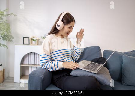 A young woman sitting on a sofa with headphones, smiling and gesturing happily while using a laptop. She looks excited and engaged, enjoying a joyful Stock Photo