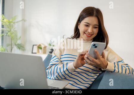 A young woman sits comfortably on the sofa, smiling while using her smartphone with a laptop in front of her, representing digital lifestyle, connecti Stock Photo