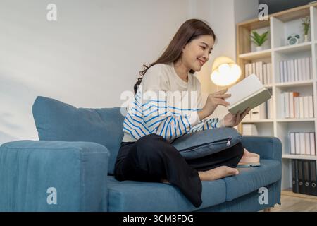 A young woman sitting on a sofa at home, relaxing with a book in her hands. She looks calm and content while enjoying reading, representing leisure ti Stock Photo
