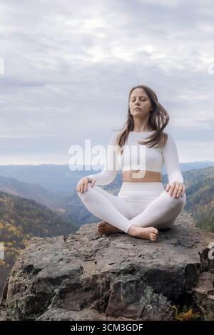 Meditating woman in lotus position on mountain cliff during autumn surrounded by nature. A woman in white relaxes on a mountaintop - outdoor sports in Stock Photo