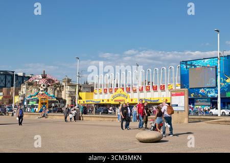 Outdoor shot of a seaside boardwalk with amusement park buildings and people, under a bright, partly cloudy blue sky. Stock Photo