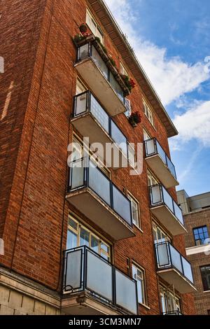 An upward-angled view of a brick building with multiple balconies, featuring frosted glass panels and metal railings, under a cloudy blue sky  in Lond Stock Photo