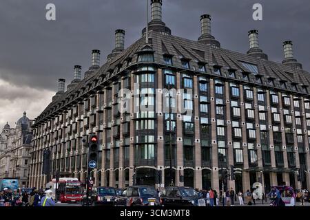 A grand, multi-story building with numerous windows and distinctive chimneys dominates the frame, under a cloudy sky. Traffic and pedestrians are visi Stock Photo