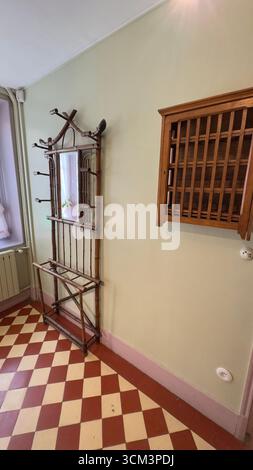A vintage bamboo coat rack with a mirror stands on a checkered floor beside a window. A wooden shelf is mounted on a pale wall. Stock Photo