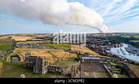 Aerial view of Whitby Abbey ruins and harbor, with a large smoke plume rising in the distance over a town. Stock Photo
