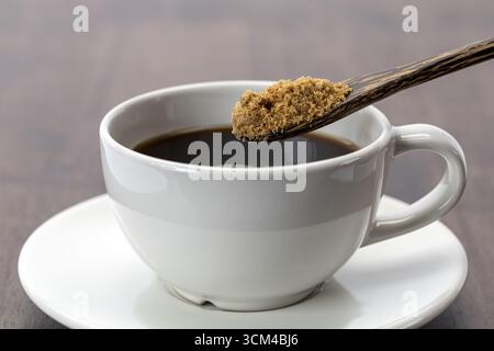 A black coffee with full brown sugar spoon, white ceramic cup and saucer on wooden table, morning beverage, warm and inviting atmosphere Stock Photo
