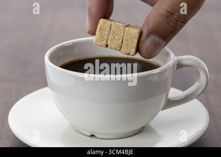 A hand adding three organic sugar cubes into cup of black coffee, creating cozy and inviting atmosphere on wooden table with white ceramic cup and sau Stock Photo