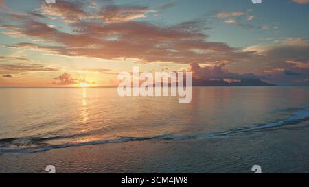 Drone fly along ocean beach coastline waves. Golden sunset colorful cloudy sky over Moorea island. Sun glow reflecting calm water shore in French Polynesia. Remote wild nature paradise, summer travel Stock Photo