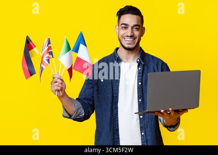 Cheerful arab guy holding laptop and international flags on yellow Stock Photo