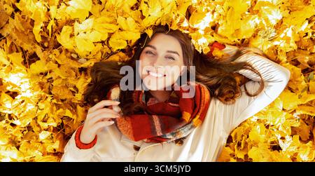Gorgeous young woman lying among yellow autumn leaves, looking at camera and smiling, top view Stock Photo