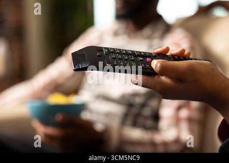 Closeup of female hand with thumb on black remote pointed at television, adjusting volume. African American couple sitting together watching tv shows or movies, relaxing in modern living room. Stock Photo