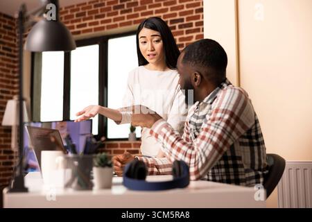 Asian woman and black man review coding work on laptop together in apartment. Young diverse couple work from home, collaborating on web app development, creating prototype for startup tech company. Stock Photo