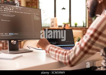 African american tech freelancer programming secure systems and reviewing coding scripts in apartment. Black male IT specialist working with multiple devices in modern home office setup. Stock Photo