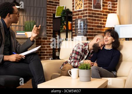 Caucasian couple hugs and laughs on couch during therapy session, sharing reconciliation moment. Boyfriend and girlfriend holding hands, receiving positive feedback from professional counselor. Stock Photo