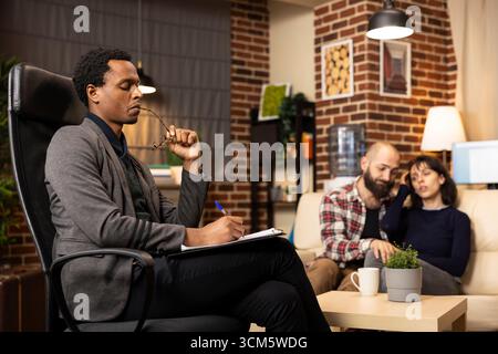 Focused male psychiatrist bites glasses and writes on clipboard while attentively listening to caucasian couple during counseling. Professional therapist engaged in ongoing therapy session. Stock Photo