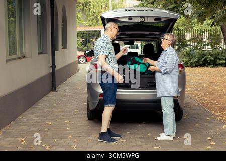Elderly people enjoying car packing preparations for their nature getaway, smiling and being excited for a comfortable road trip during their retirement. Happy relaxed lifestyle. Stock Photo