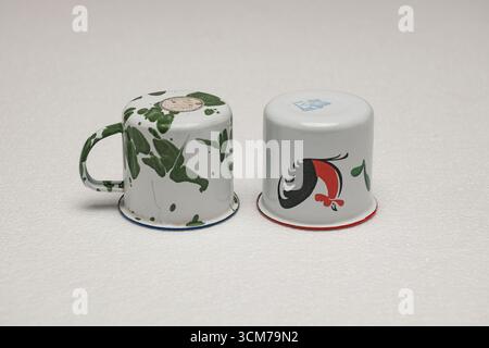 Two vintage enamel cups, one with green splatter paint and the other with a red and black design, placed on a white surface Stock Photo