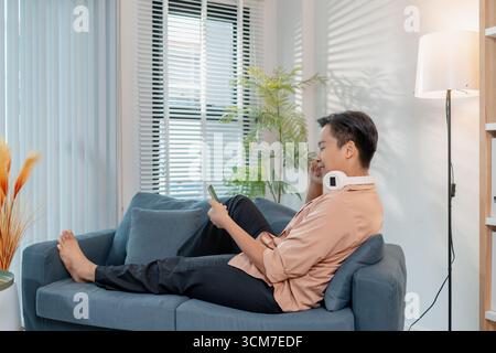 A young man relaxes at home, smiling while using his smartphone. He enjoys leisure time with headphones around his neck, sitting comfortably and feeli Stock Photo