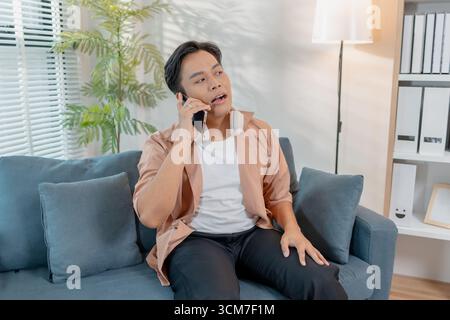 A young man sitting on a sofa with headphones around his neck is talking on the phone, smiling and gesturing playfully, showing relaxed and joyful com Stock Photo