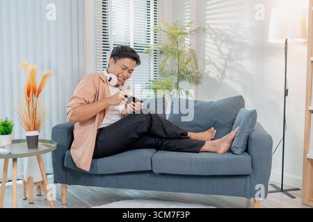 A young man sits on a sofa at home, wearing casual clothes with headphones around his neck, smiling and celebrating while looking at his smartphone, s Stock Photo