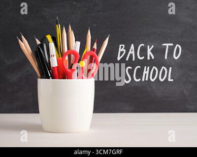 Pencils, pens, scissors and other school supplies in a white container on a desk in front of a blackboard with the words back to school written on it Stock Photo