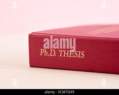 Close-up of a hardcover PhD thesis book with a red cover on a wooden table. PhD thesis and graduation concept. Stock Photo