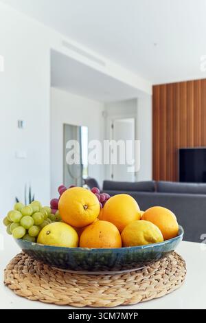 Colorful ceramic bowl filled with oranges, lemons, and grapes placed on woven placemat atop modern white dining table Stock Photo