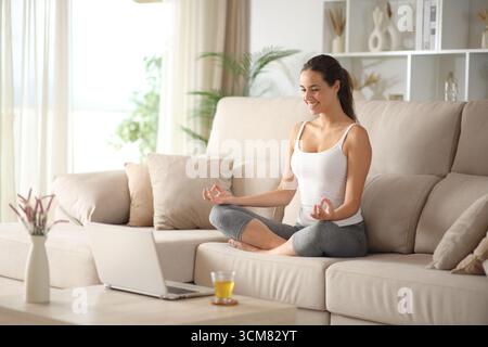 Happy yogi e-learning yoga online with a laptop sitting on a couch at home Stock Photo