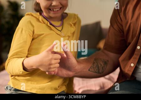 Caucasian child girl smiling while playing thumb wrestling game with adult Caucasian man, both hands visible with focus on interaction, man showing tattooed forearm, girl wearing necklace Stock Photo