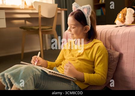Caucasian girl child sitting on floor drawing in sketchbook, wearing animal ear headband, smiling and looking to side, bedroom setting with desk and bed in background Stock Photo