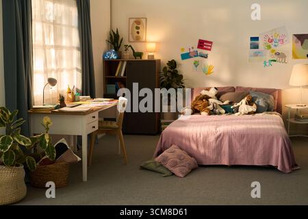 Child bedroom interior featuring neatly made bed with plush toys, study desk with school supplies, wall decorated with colorful drawings, large window letting in natural light Stock Photo