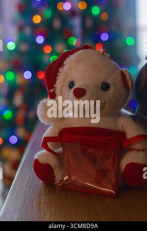 Adorable white teddy bear in Santa hat sitting near Christmas tree with colorful lights and festive bokeh. Cozy holiday mood and warmth Stock Photo