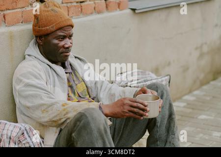 Homeless Black Man Sitting Against Wall Holding Cup Appearing Contemplative Stock Photo