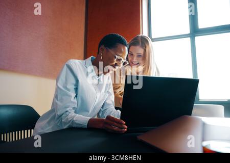 Two women smiling and collaborating on a laptop in a modern office. The scene captures teamwork, professionalism, and a sense of positive working envi Stock Photo
