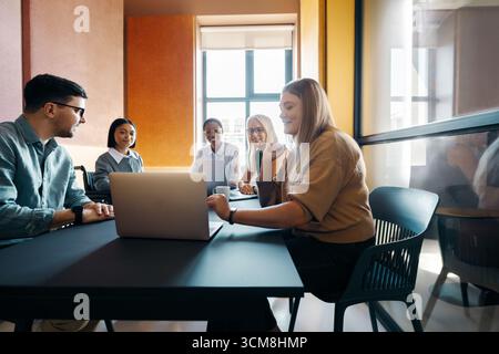Group of young professionals, both men and women, working together at a shared table in a brightly lit, contemporary office environment. They share id Stock Photo