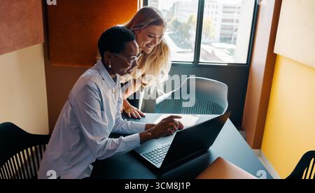 Two professional women collaborate in a modern office space, discussing ideas while using a laptop. Their teamwork exhibits creativity, innovation, an Stock Photo
