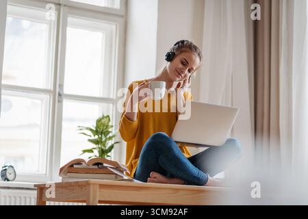 Young female student sitting at the table, using headphones while studying. Stock Photo