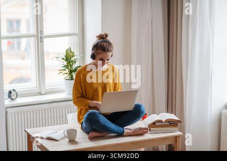 Young female student sitting at the table, using headphones while studying. Stock Photo