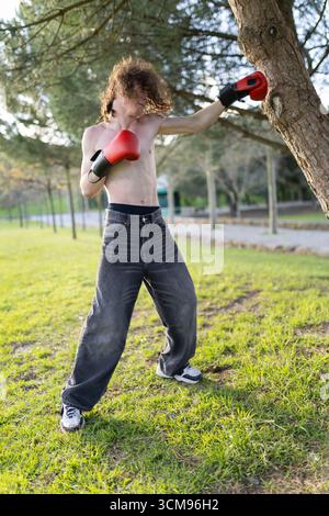 Young man practicing boxing in park, punching tree with boxing gloves Stock Photo