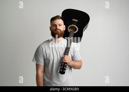 Bearded young man in grey t-shirt holding black acoustic guitar on shoulder in studio Stock Photo