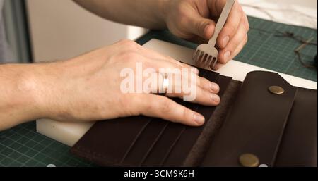 Punching stitching holes in brown leather wallet with pricking iron in artisan workshop Stock Photo