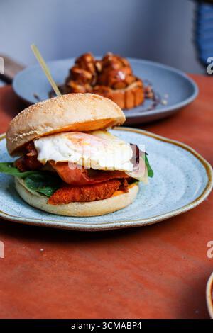 breakfast sandwich served on a rustic plate, set in a cozy cafe environment. The substantial sandwich is built on a toasted sesame seed bun Stock Photo