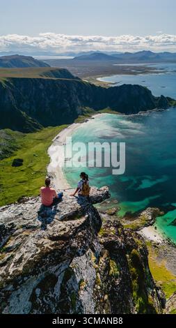 Two adventurers sit atop Matind Mountain, taking in the stunning panoramic views of Bleiks coastline and turquoise waters. This serene landscape showcases Norways natural beauty on a sunny day. Stock Photo