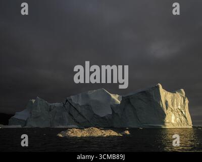 Large iceberg illuminated by sunset light under dark clouds in ocean near Disko Bay, Greenland Stock Photo