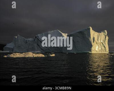 Large iceberg illuminated by sunset light under dark clouds in ocean near Disko Bay, Greenland Stock Photo