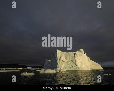 Large iceberg illuminated by sunset light under dark clouds in ocean near Disko Bay, Greenland Stock Photo