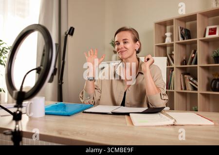 A professional woman engages in an online meeting using a ring light and laptop in her home office. She appears confident and enthusiastic, surrounded Stock Photo